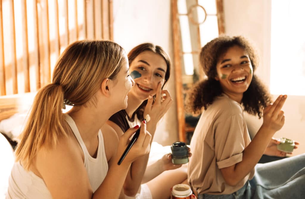 Three charming teenagers of different races apply cosmetic masks to their faces in the bedroom. Female multiethnic friends have fun together at home.